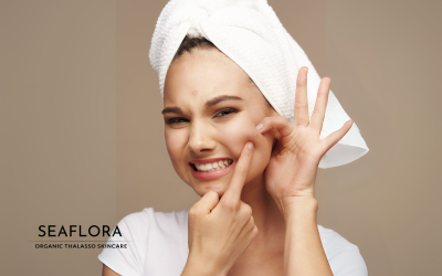A woman with a towel wrapped around her head and a white T-shirt makes a funny face as she tries to pop a pimple, with the Seaflora Skincare logo in the corner.