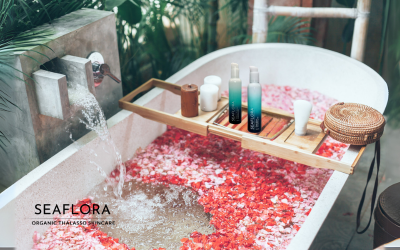 A serene bath filled with pink flower petals, with a wooden bath table holding Seaflora’s Everything Oils and the Seaflora Skincare logo displayed.