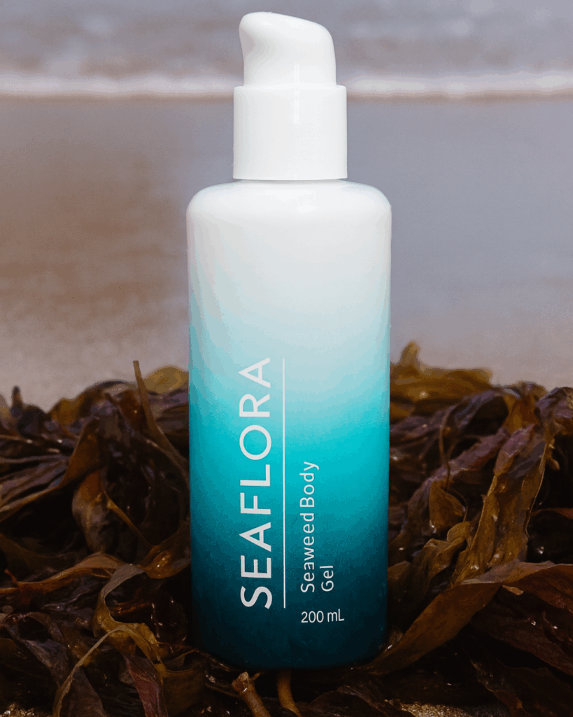 Seaweed Body Gel by the Pacific — Nature Meets Skincare - Seaflora Skincare A high-resolution photo of Seaflora’s Seaweed Body Gel standing upright on a textured driftwood surface, with vibrant green strands of hand-harvested wild seaweed resting beside it. The bottle’s gradient blue design evokes the ocean, emphasizing the product’s raw, organic, seaweed-based formulation.