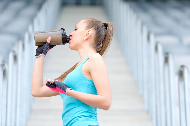 Active Woman Hydrating with Protein Shake Outdoors - Seaflora Skincare A woman in athletic gym wear sits on outdoor concrete steps, taking a break and drinking from a protein shake bottle to combat aging skin on a sunny day.