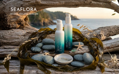 A rustic-luxury still life of Seaflora products on a Vancouver Island beach. Two white-to-teal gradient bottles, Sea Splash Toning Essence and Deep Sea Facial Moisturizer, stand on weathered driftwood surrounded by smooth grey sea stones and long, dark bull kelp fronds. In the foreground, a small ceramic dish holds a dollop of cream with an applicator brush. The scene is framed by a natural driftwood arch, revealing a soft golden hour view of the Pacific Ocean and distant mountains.